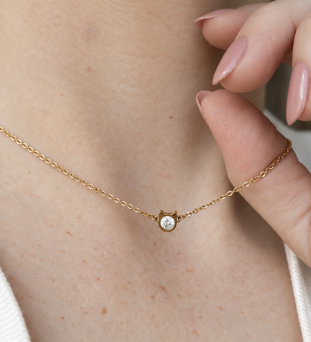 A close-up lifestyle shot of a delicate gold chain necklace worn around a person's neck, featuring a small pendant shaped as a little cat head, with a zirconia stone in the middle. The person's hand with manicured nails gently rests near the necklace.