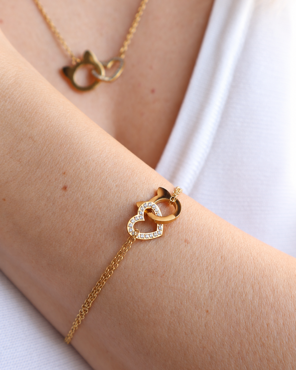 A gold chain bracelet with an interlocking double-heart pendant, worn on a person's wrist. The bracelet features one heart with small clear crystals and one solid gold cat shape.