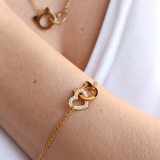 A gold chain bracelet with an interlocking double-heart pendant, worn on a person's wrist. The bracelet features one heart with small clear crystals and one solid gold cat shape.
