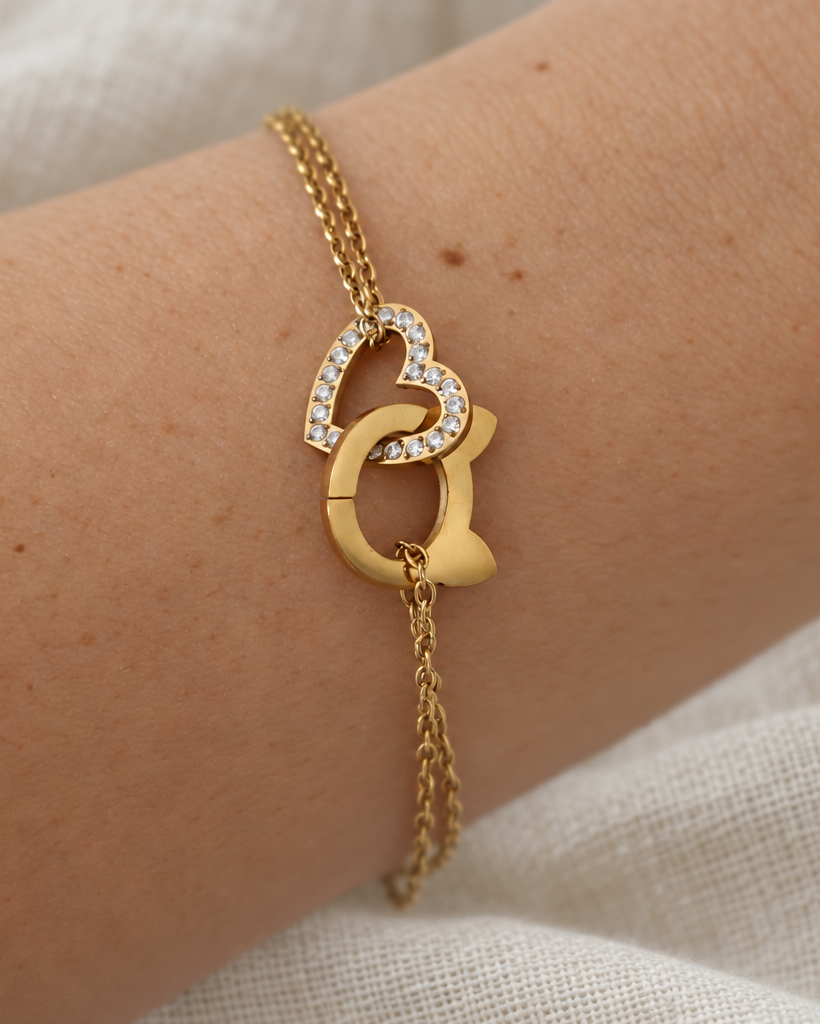 A gold chain bracelet with interlocking heart and cat head charms, one of which is set with small clear stones, shown worn on a person's wrist against a soft beige fabric background.