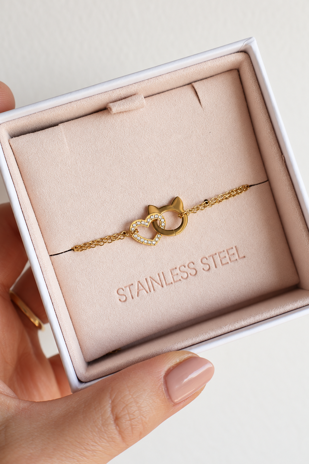 A gold-toned stainless steel bracelet with two interlocked pendants, one pendant is shaped as a cat head, and other one is heart paved with zirconia, displayed inside a light pink jewellery box held by a hand.