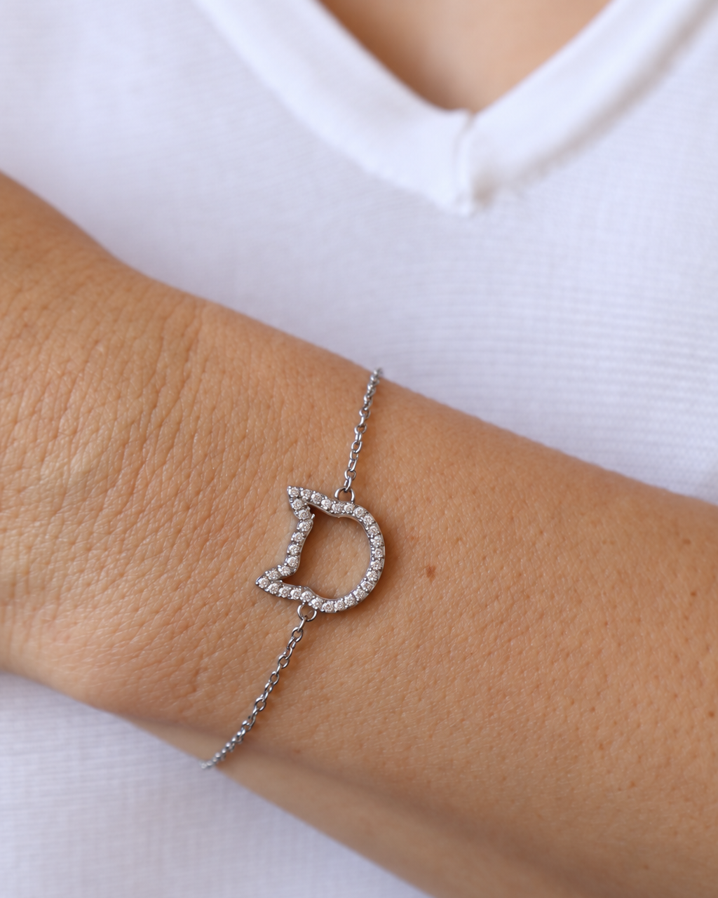 A stainless steel chain bracelet with a cat head charm paved with small zircon stones, shown worn on a person's wrist against a white shirt background.
