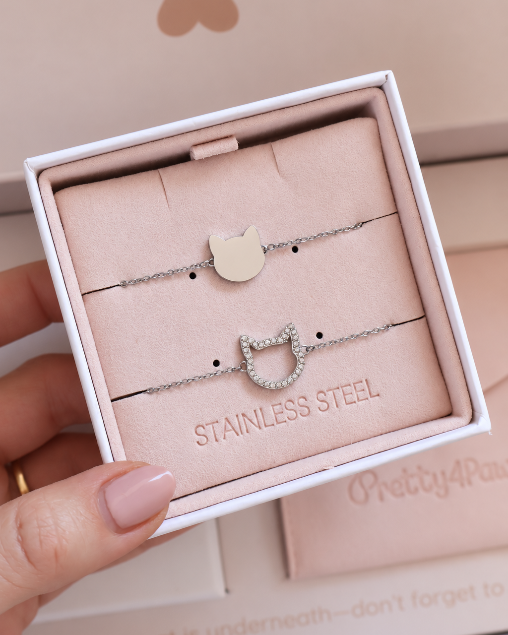 A hand holds an open white jewelry box containing two delicate stainless steel cat-themed bracelets on a blush pink insert. One bracelet has a smooth silver-toned cat head charm, and the other has a cat head outline encrusted with small zirconia stones.