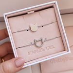 A hand holds an open white jewelry box containing two delicate stainless steel cat-themed bracelets on a blush pink insert. One bracelet has a smooth silver-toned cat head charm, and the other has a cat head outline encrusted with small zirconia stones.